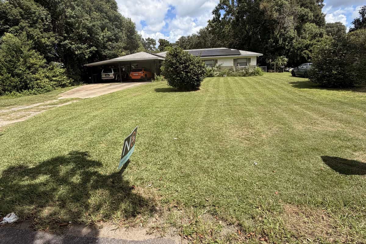 A wide, freshly mowed front lawn in front of a single-story home with a carport, with mature trees behind
